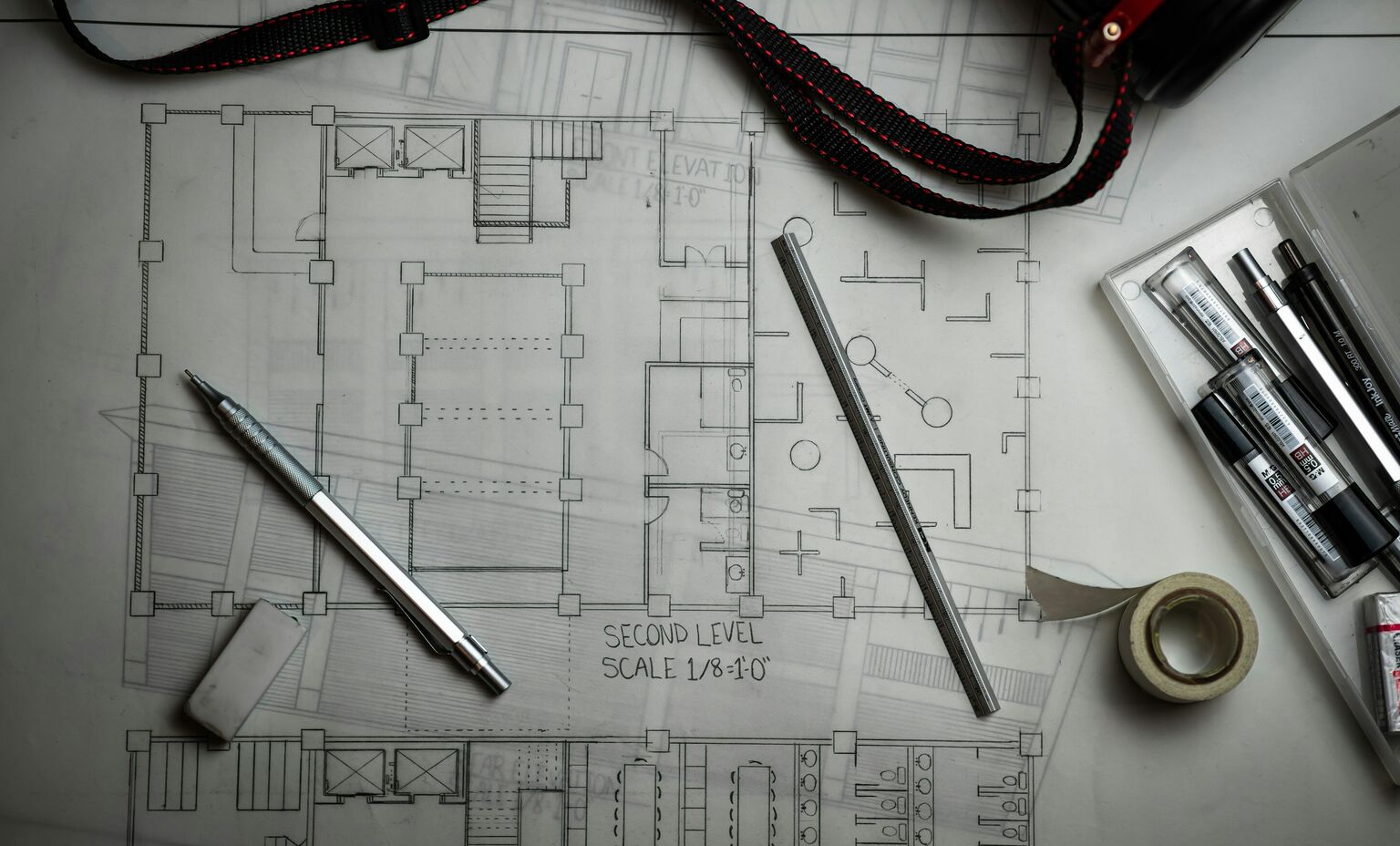 Illustrative image: an architectural floor plan on a desk with a mechanical pencil, a ruler, an eraser, masking tape and a box of pens